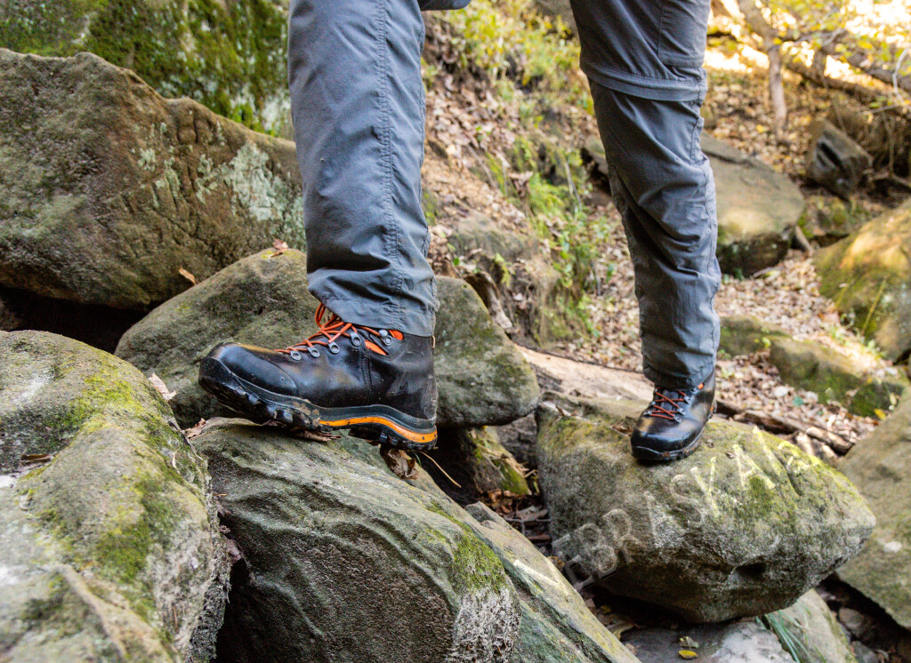 Hiker with gray pants stepping on mossy rocks in a wooded area.