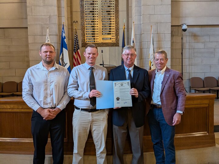 Four men stand inside a formal hall holding a certificate; flags and ornate walls in the background.